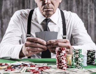 Poker player. Close-up of serious senior man in shirt and suspenders sitting at the poker table and holding cards with money and gambling chips laying all around him